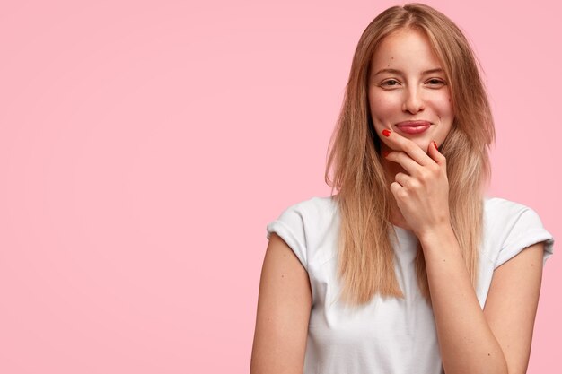 happy European woman holds chin, looks positively, hears pleasant strory from friend, dressed in casual outfit