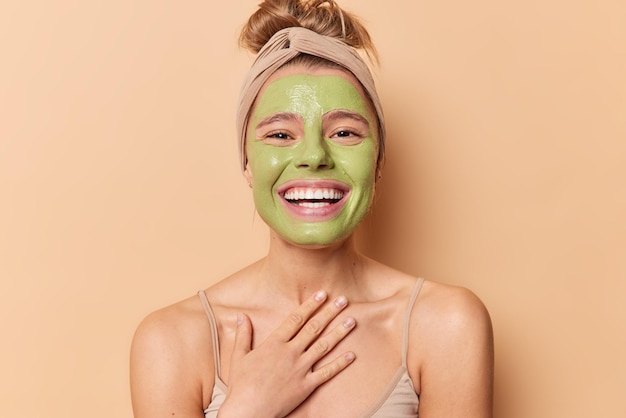 Счастливая европейская женщина широко улыбается, держит руку на груди, наносит питательную зеленую маску, проходит косметические процедуры, носит повязку на голову, расчесывает волосы, изолированные на коричневом фоне Концепция ухода за кожей