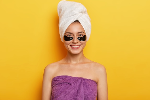 Счастливая европейская женщина с нежной улыбкой, имеет черные пятна коллагена, уменьшает проблему темных кругов под глазами, завернута в полотенце на голове и по всему телу, улучшает состояние кожи.