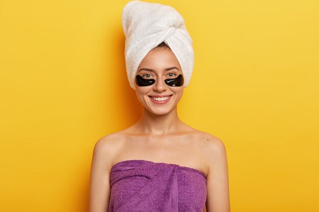 Happy European woman with gentle smile, has black collagen patches, reduces problem of dark circles under eyes, wrapped in towel on head and over body, improves skin condition