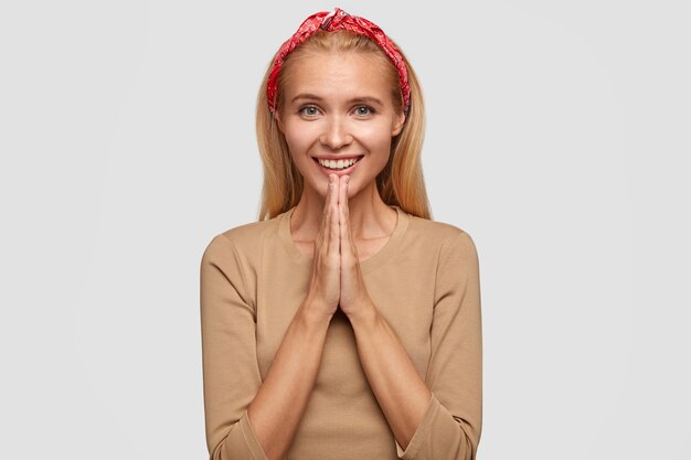 Happy European young female with blonde hair, keeps hands in praying gesture