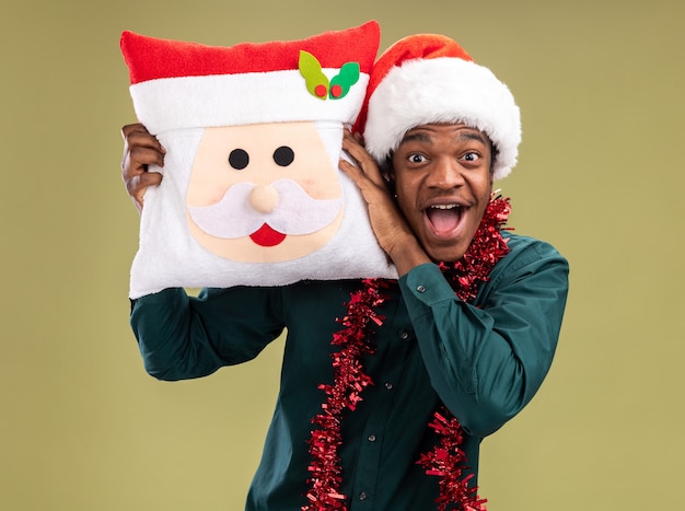 녹색 배경 위에 서있는 카메라를보고 크리스마스 베개를 들고 갈 랜드와 산타 모자에 행복하고 흥분 아프리카 계 미국인 남자