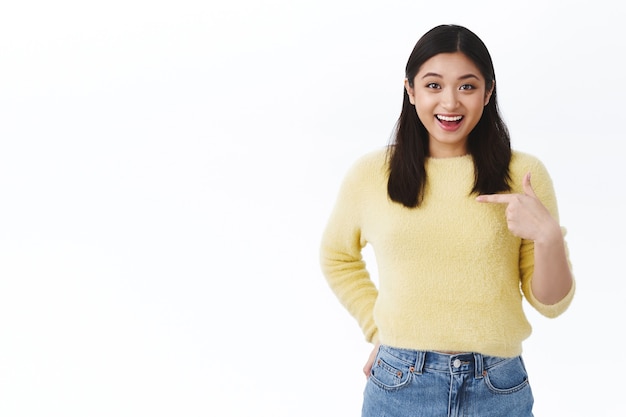 Happy excited asian girl pointing at herself surprise as being told good news, winning awesome prize, smiling upbeat, feeling rejoice and satisfaction of achieving success, standing white wall