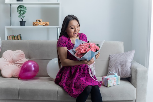 Happy and excited asian young woman in beautiful dress smiling cheerfully sitting on a couch with bouquet of flowers and present in light living room celebrating international women's day