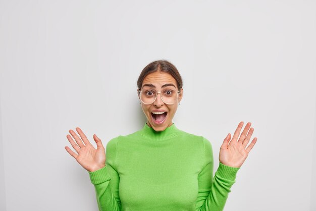 Happy excited brunette European woman exclaims loudly keeps palms raised gets unexpected news wears transparent glasses and green turtleneck isolated over white background. Human reactions concept