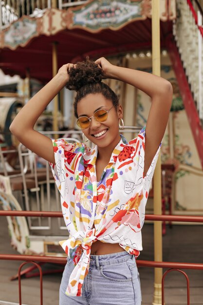 Happy excited curly brunette woman in denim pants, stylish cropped colorful blouse and orange sunglasses touches hair, smiles and poses near carousel
