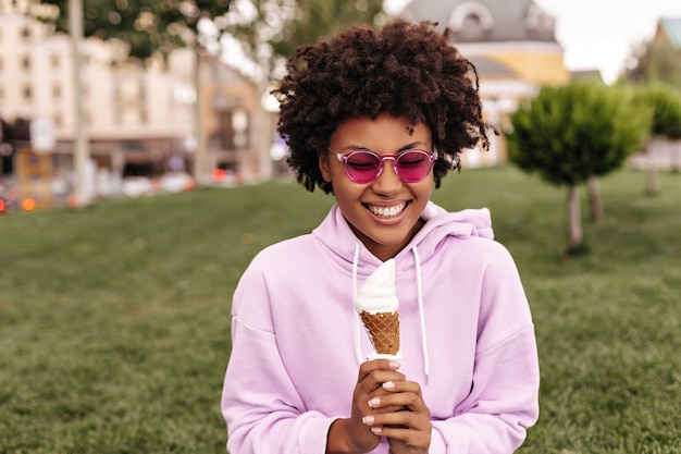 Happy excited curly woman in fuchsia sunglasses and pink hoodie smiles sincerely and holds ice-cream