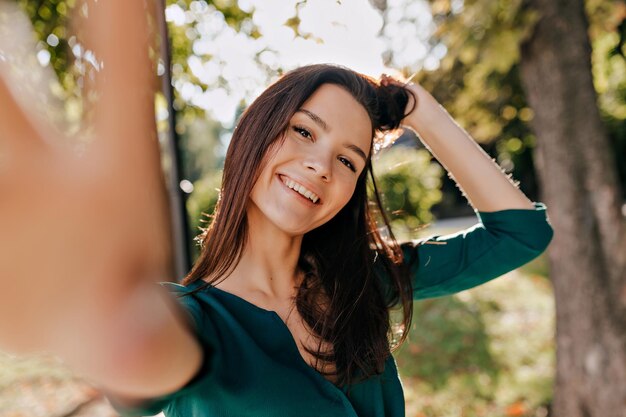 Счастливая взволнованная темноволосая женщина с прекрасной улыбкой в зеленой блузке делает селфи в солнечном городском парке