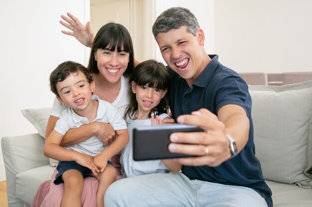 Happy excited family couple hugging adorable kids, sitting on couch at home together, taking selfie on phone.
