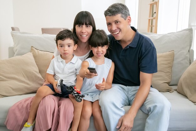 Happy excited family couple and two kids watching TV together, sitting on couch in living room, using remote control.