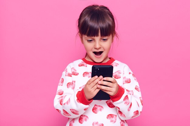 Счастливый взволнованный ребенок женского пола в перемычке с сердечками, держа в руках сотовый телефон и видит что-то удивительное на его экране, держит рот открытым, позируя изолированно над розовой стеной.