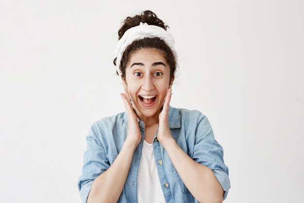 Happy excited female has dark hair, wearing do-rag and denim shirt, glad to see something unexpected, keeps hands on cheeks, smiles broadly. Overjoyed woman stares  with joyful expression