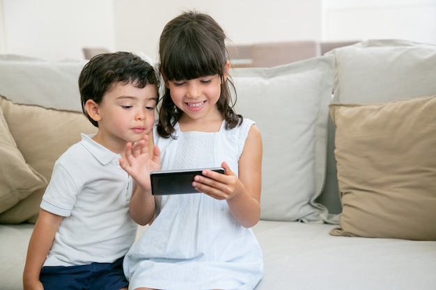 Happy excited girl and her little brother playing online game on phone while sitting on couch in living room.