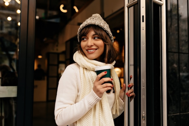 Happy excited girl in white knitted clothes came out of cafe with coffee High quality photo