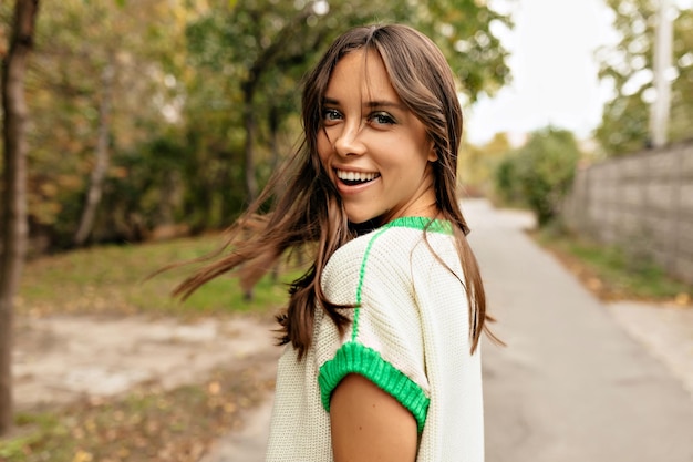 Free photo happy excited girl with flying darkhaired girl is turn around at camera while walking down on the road with green and yellow trees happy girl walking in the city