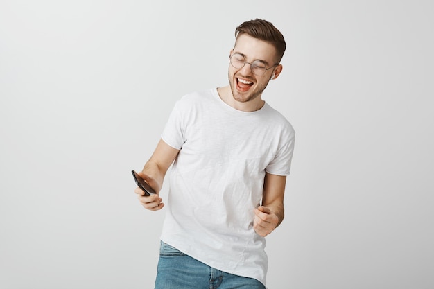 Happy excited guy smiling, looking at mobile phone and dancing in wireless earphones