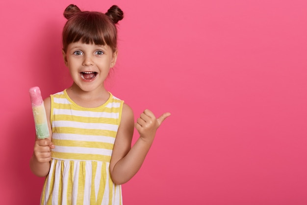 Happy excited little kid with ice cream with widely opened mouth, pointing thumb aside, funny girl with knots, copy space for advertisement.