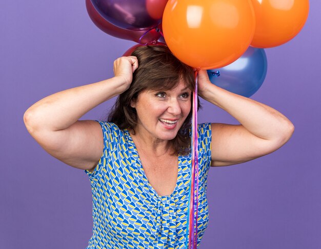 Счастливая и взволнованная женщина среднего возраста, держащая кучу разноцветных шаров, празднует день рождения, стоя над фиолетовой стеной