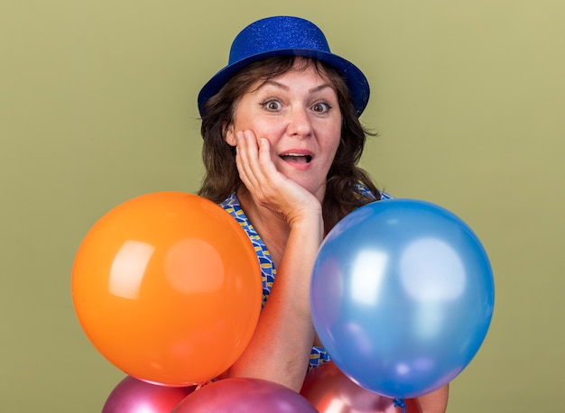 Happy and excited middle age woman in party hat with bunch of colorful balloons