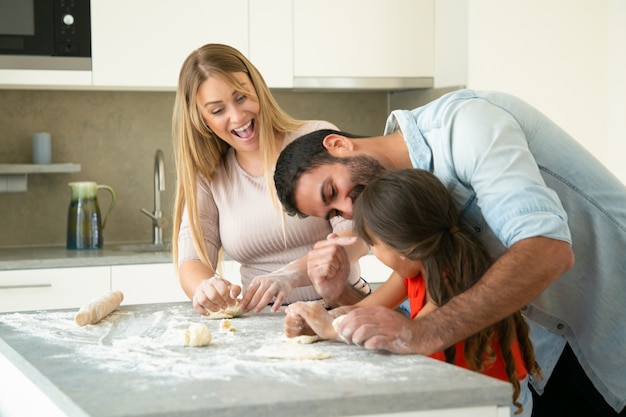 Happy excited mom and dad having fun while teaching daughter to make dough at kitchen table. Young couple and their girl baking buns or pies together. Family cooking concept