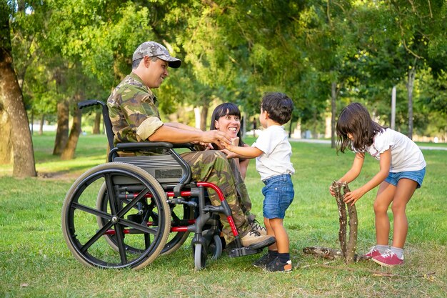 幸せな興奮したお母さんと車椅子の障害者の軍のお父さんは、屋外で子供たちと余暇を過ごし、芝生の上で火のために薪を配置します。傷痍軍人または家族の屋外の概念