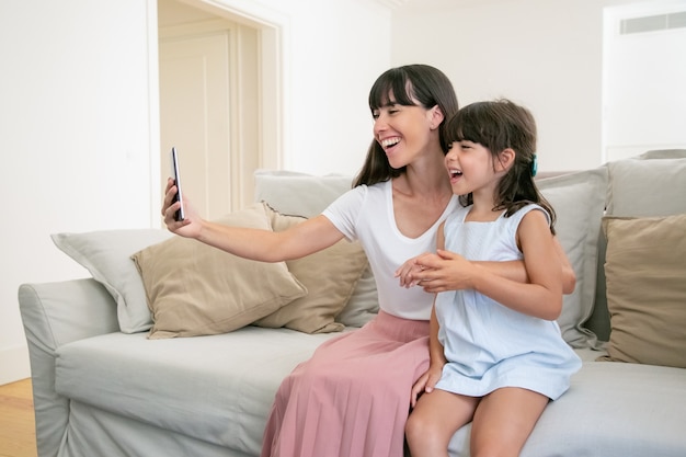 Happy excited mom and little daughter using phone for video call while sitting on sofa at home together