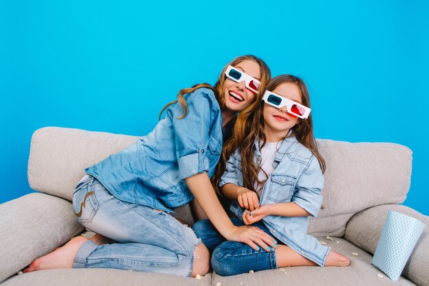 Счастливая взволнованная мать с милой красивой дочерью на диване на синем фоне. Вместе смотреть 3D-фильм в очках, носить джинсовую одежду, выражать позитив и счастье перед камерой