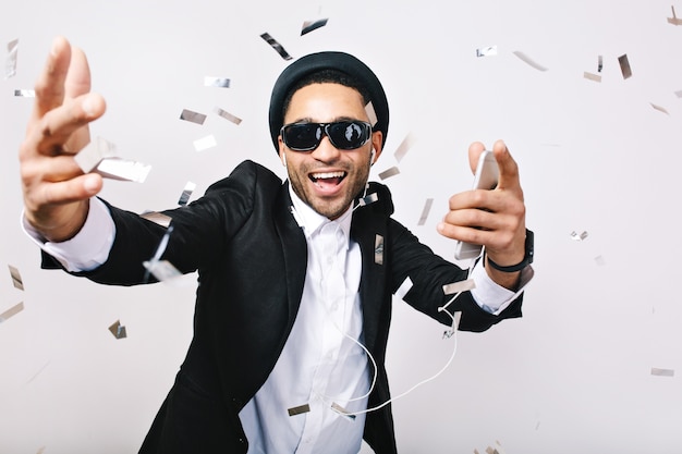 Happy excited party time of joyful handsome guy in hat, suit, black sunglasses having fun in tinsels. Listening to music through headphones, celebrating, singer, super star.