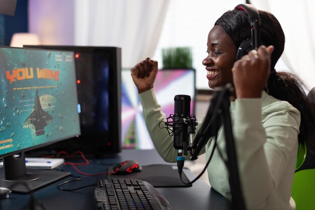 우주 슈팅 게임 대회에서 우승한 후 손을 드는 행복한 흥분된 프로 게이머 여성. RBG 컴퓨터 장비를 사용하여 온라인 비디오 게임을 하는 성공적인 승자. 홈 스튜디오에서. 사이버 공간
