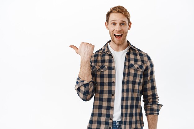 Happy and excited redhead man pointing finger left at awesome new promo offer showing advertisement banner with amazed smiling face standing over white background