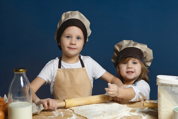 Счастливый взволнованный школьник раскладывает тесто скалкой, пока его младшая сестра помогает ему. Две милые дети, братья и сестры вместе готовят пиццу