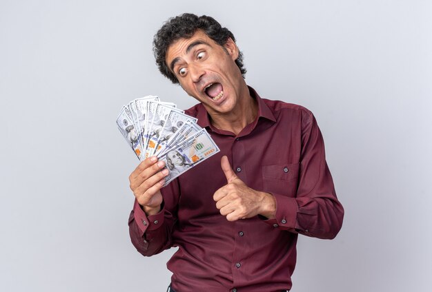 Happy and excited senior man in purple shirt holding cash looking at money smiling cheerfully showing thumbs up standing over white background