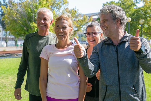 Бесплатное фото Счастливые взволнованные спортивные зрелые люди, стоящие вместе после утренней зарядки в парке, глядя в сторону и улыбаясь, делая жест пальца вверх. концепция выхода на пенсию или активного образа жизни