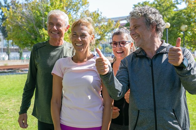 Happy excited sporty mature people standing together after morning exercises in park, looking away and smiling, making thumb up gesture. Retirement or active lifestyle concept