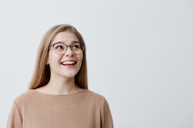 happy excited surprised female model with blonde straight hair in glasses, being pleased with something, can`t believe in her success, glad to be praised, poses against gray studio background