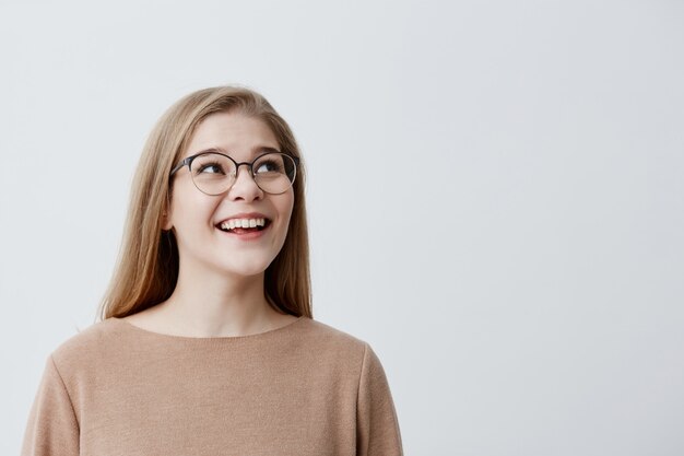 Happy excited surprised female model with blonde straight hair in glasses, being pleased with something, can`t believe in her success, glad to be praised, poses against gray studio background