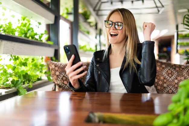 Happy excited woman receiving text message with news about discounts in shop on mobile phone sitting in cafe with loft interior, woman holding smartphone with shocked surprised expression