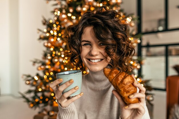 Счастливая возбужденная женщина с короткими вьющимися волосами позирует с круассаном и кофе рождественской елки