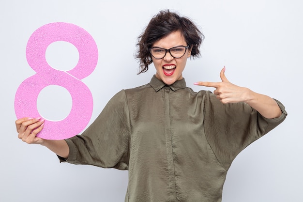Free photo happy and excited woman with short hair holding number eight made from cardboard pointing with index finger at it celebrating international women's day march 8 standing over white background