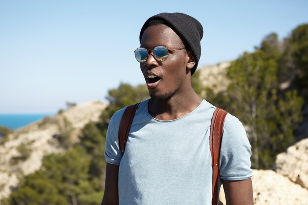 Happy excited young African American male traveler in stylish shades and headwear looking stunned, keeping mouth wide opened while standing high in mountains and admiring beautiful view below
