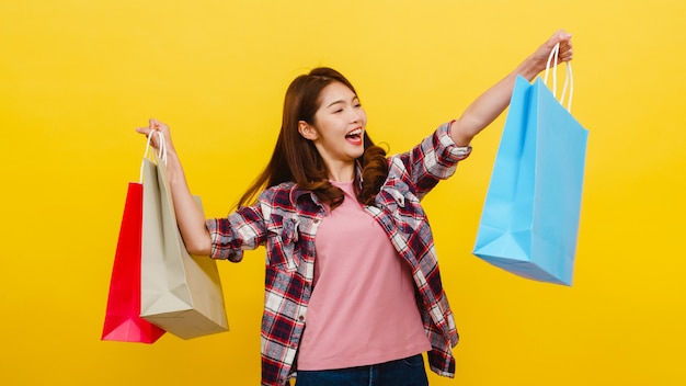 幸せな興奮してアジアの若い女性がカジュアルな服で立ち上がって黄色の壁を越えてカメラを見て手で買い物袋を運ぶします。顔の表情、季節限定セール、消費者のコンセプト。