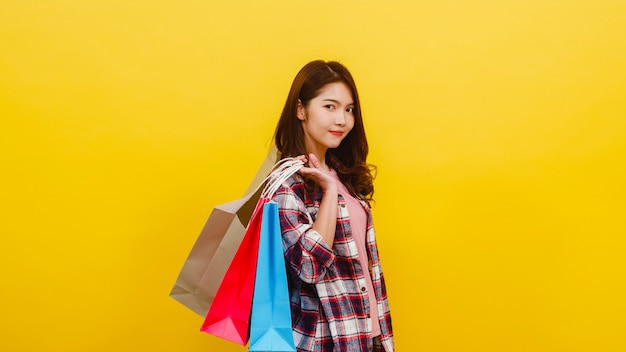 Happy excited young Asian lady carrying shopping bags with hand raising up in casual clothing and looking at camera over yellow wall. Facial expression, seasonal sale and consumerism concept.