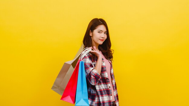 Happy excited young Asian lady carrying shopping bags with hand raising up in casual clothing and looking at camera over yellow wall. Facial expression, seasonal sale and consumerism concept.