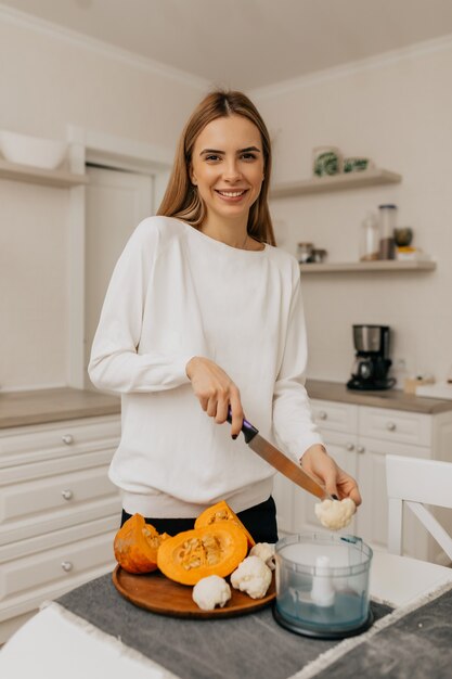 キッチンで野菜を調理しながらカメラでポーズをとって白いシャツで幸せな興奮した若い女性