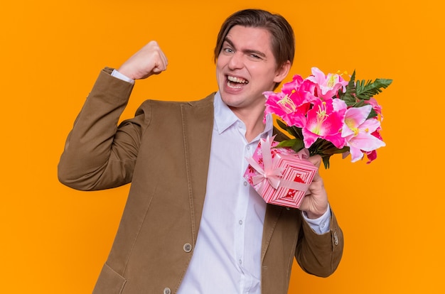 Happy and excited young man holding present and bouquet of flowers clenching fist going to congratulate with international women's day standing over orange wall
