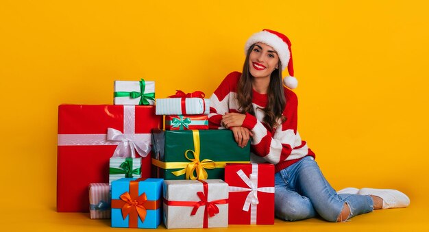 Happy excited young smiling beautiful and charming woman in santa hat and with christmas mood while she sitting with many colorful gift boxes and having fun