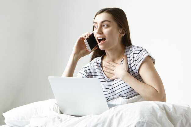happy excited young unemployed woman sitting in her bed with pc on her lap and having phone conversation with employer
