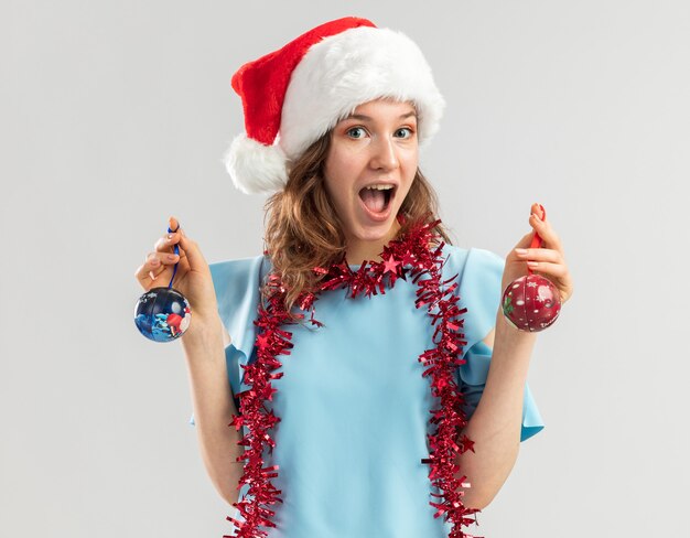 파란색 상단과 산타 모자에 행복하고 흥분된 젊은 여자가 유쾌하게 웃는 크리스마스 공을 들고 그녀의 목에 반짝이