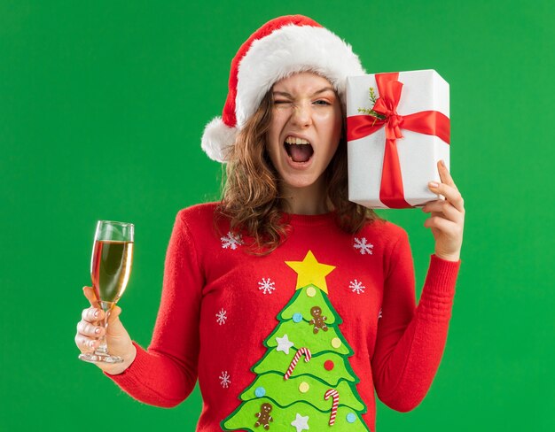 Happy and excited young woman in red christmas sweater  and santa hat holding  glass of champagne and present standing over green  background