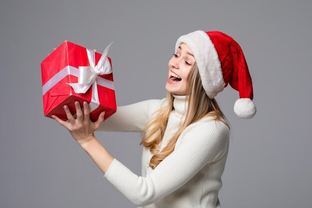 Счастливая возбужденная молодая женщина в шляпе санта-клауса с подарочной коробкой, изолированной над серой стеной